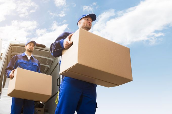 Two movers in blue coveralls unloading cardboard boxes from a truck against a blue sky.