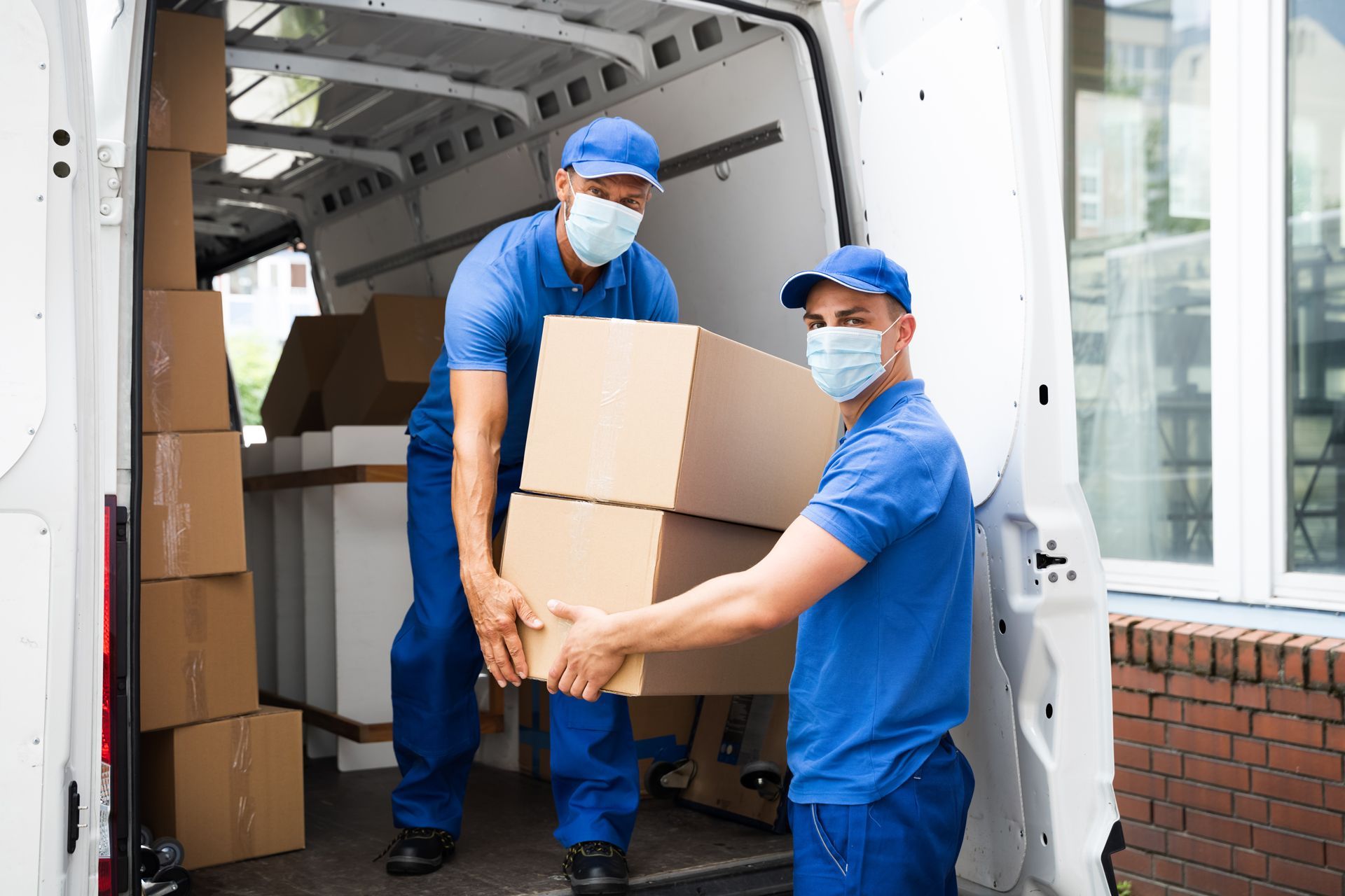 Two workers in blue uniforms and masks carrying boxes into a moving truck.