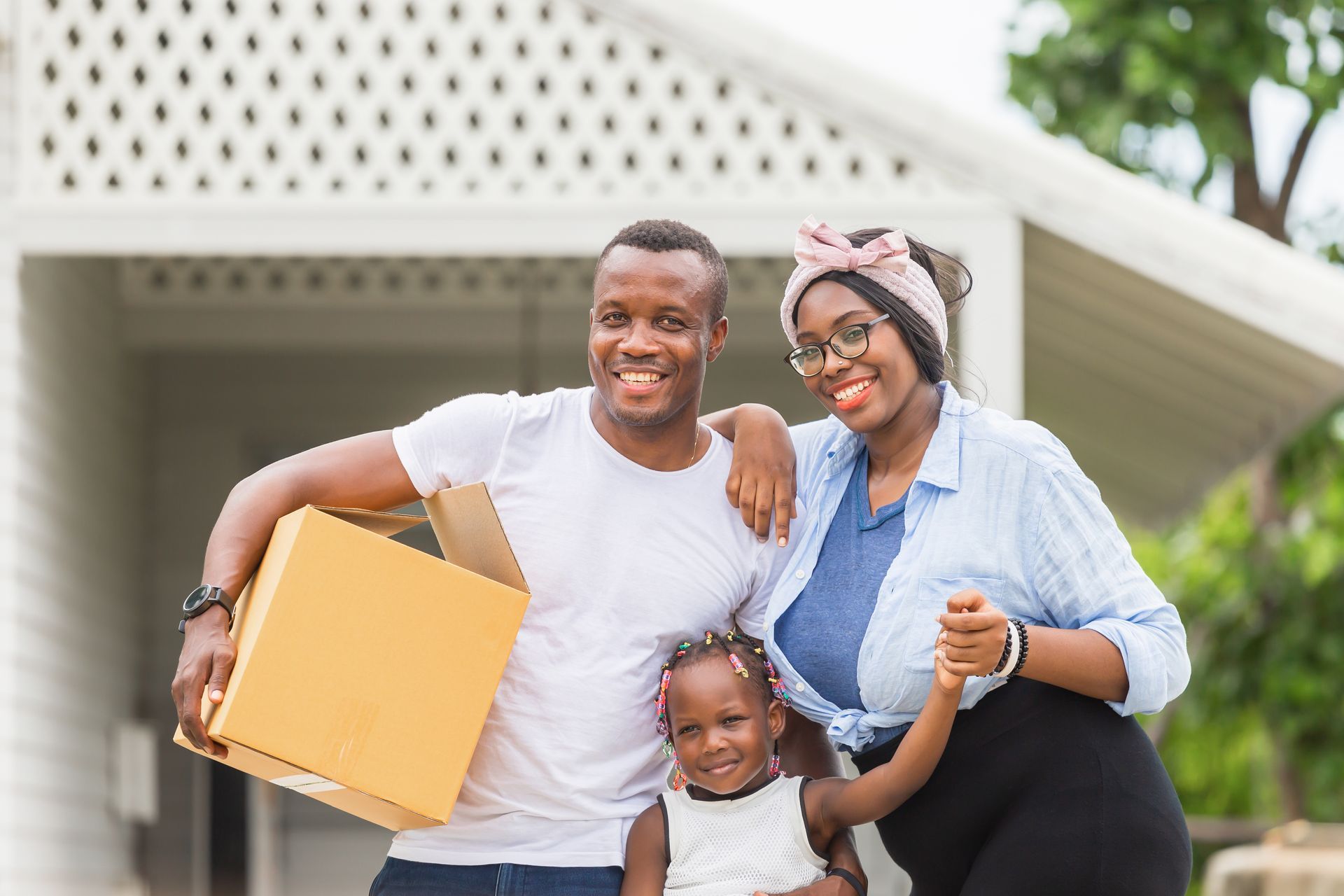 Family of three smiling in front of a white house, man holds box, woman has arm around child.