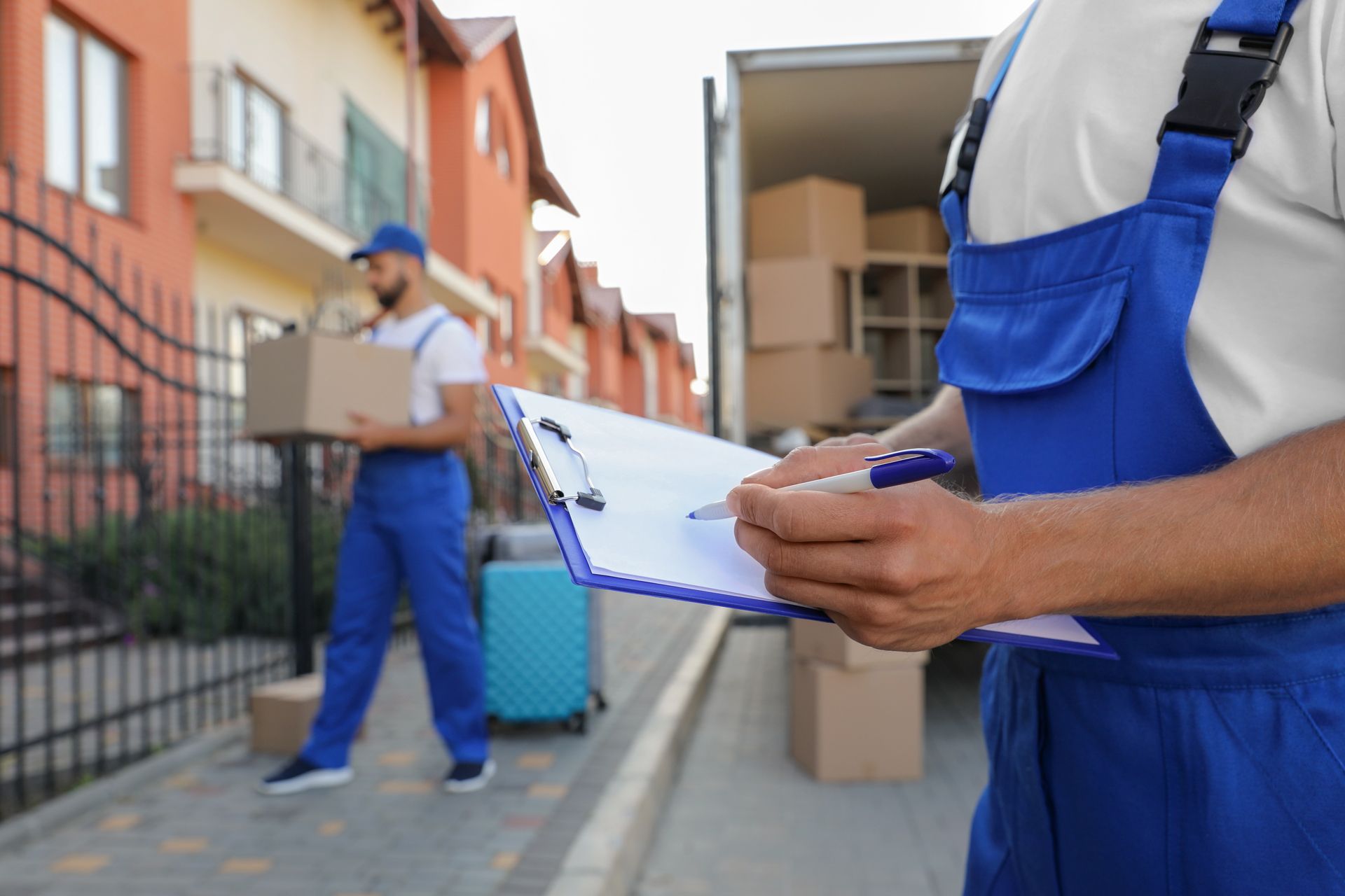 Movers in blue uniforms loading boxes from a home into a truck. One is writing on a clipboard.