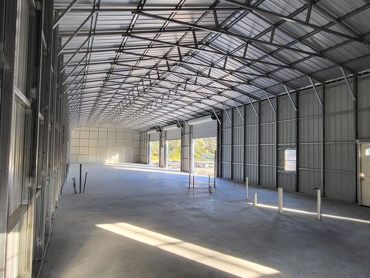 Empty warehouse interior with metal frame roof, concrete floor, and sunlight streaming in from open sides