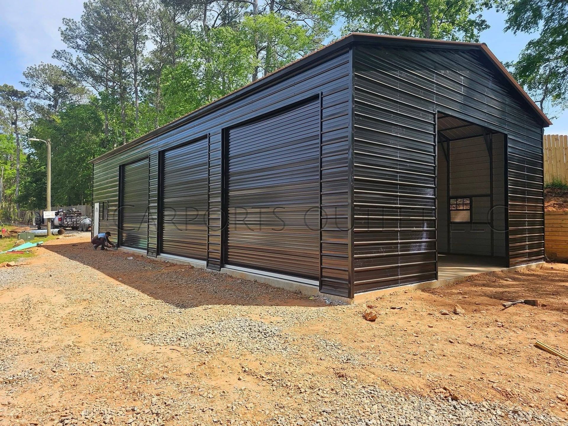 Black metal-sided building under construction on a dirt lot, with trees in the background