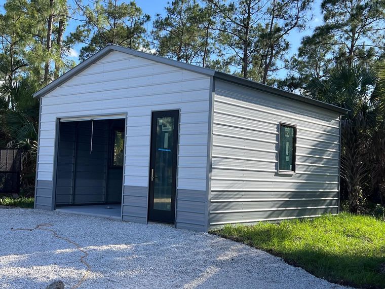 Small gray shed with open garage-style door, side door, and green window trim on a gravel driveway