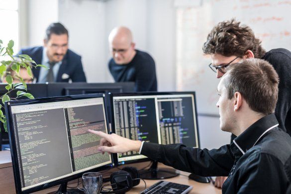 A man is pointing at a computer screen while another man looks on.