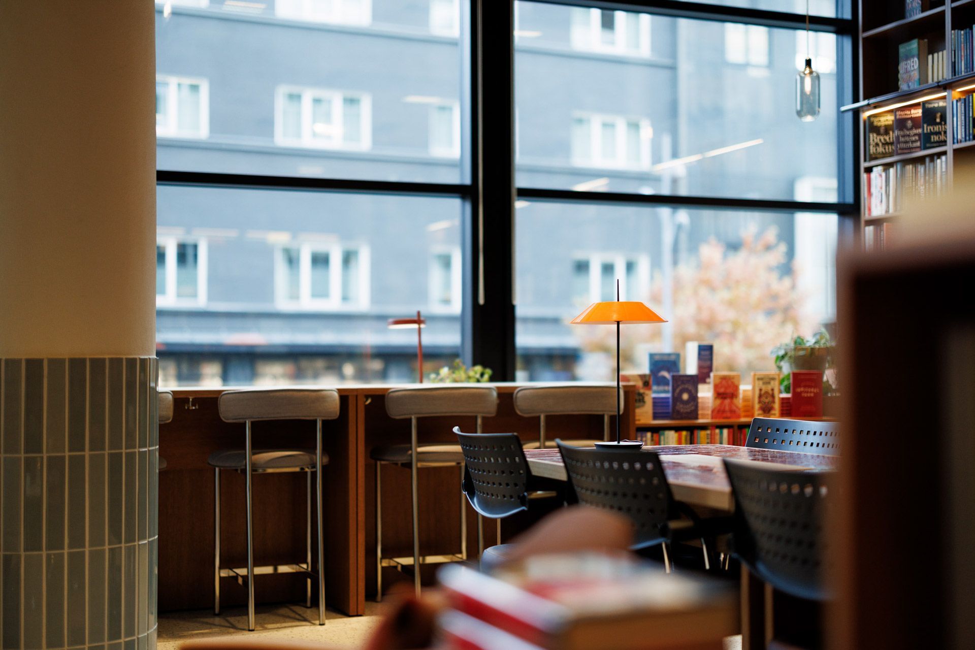 Moderne kaféinteriør med krakker, en glødende oransje lampe og store vinduer med utsikt over bygninger