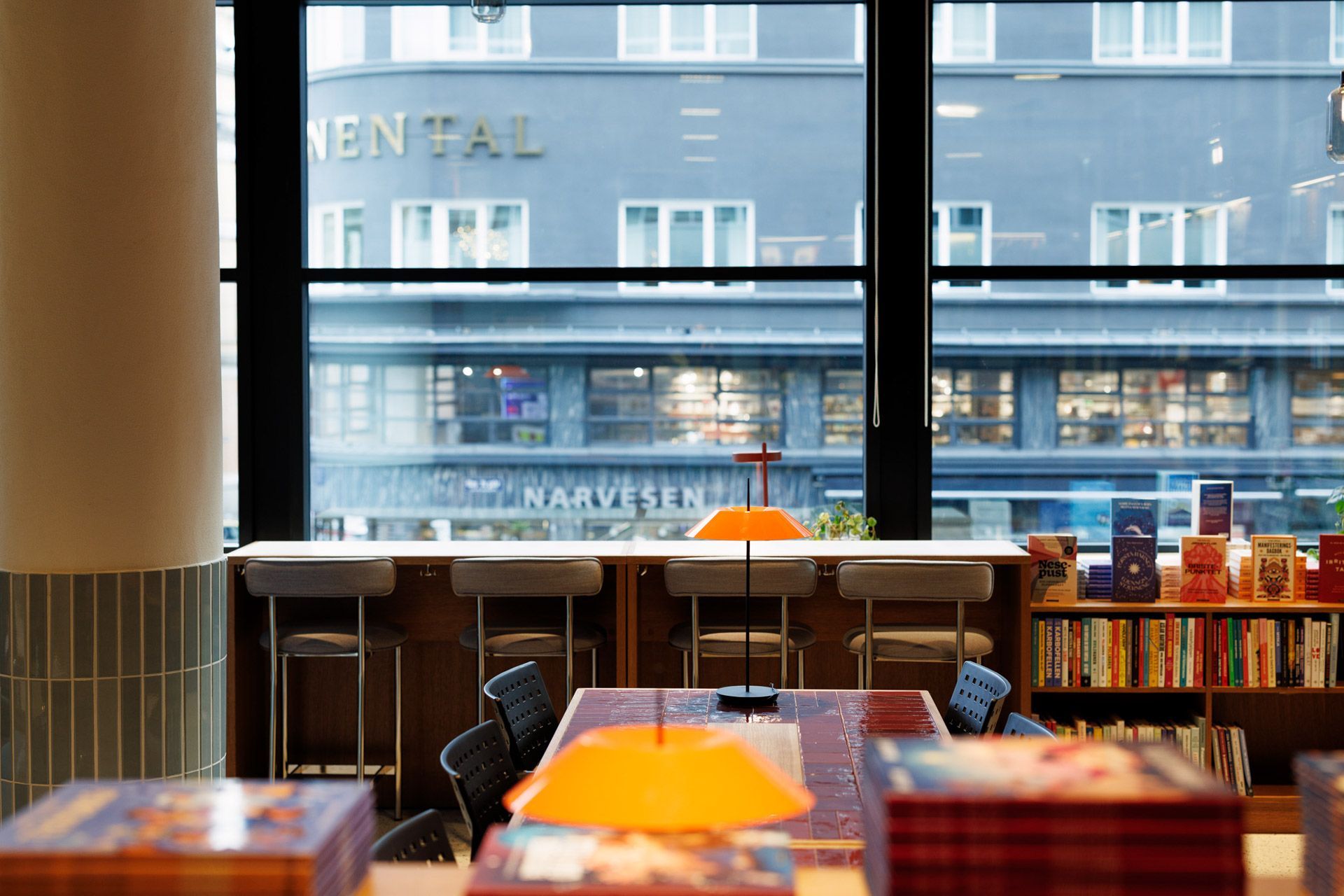 Lyst kaféinteriør med bord og oransje lamper med utsikt over en bygate gjennom store vinduer