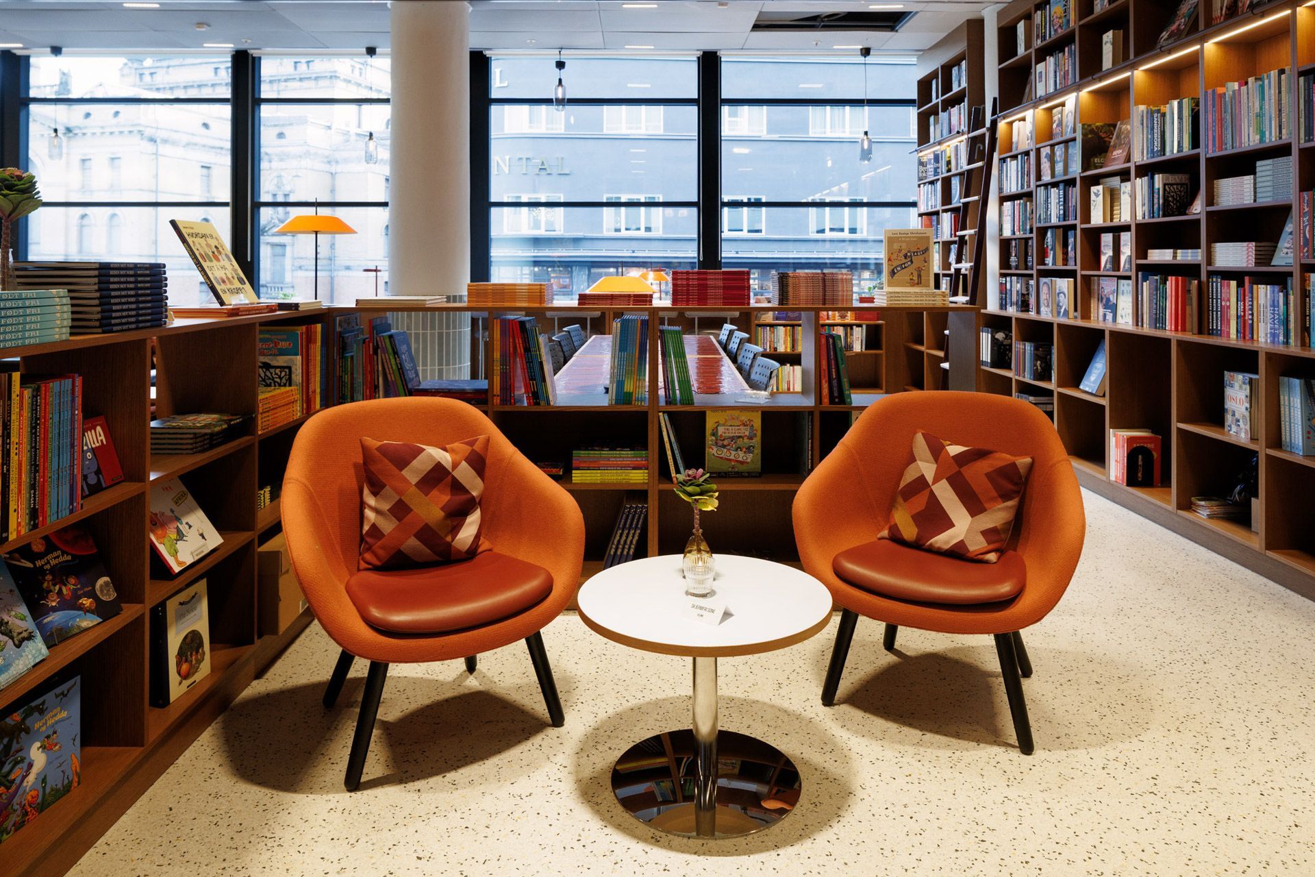 Moderne bibliotekstue med to oransje stoler, rundt hvitt bord, bokhyller og store vinduer