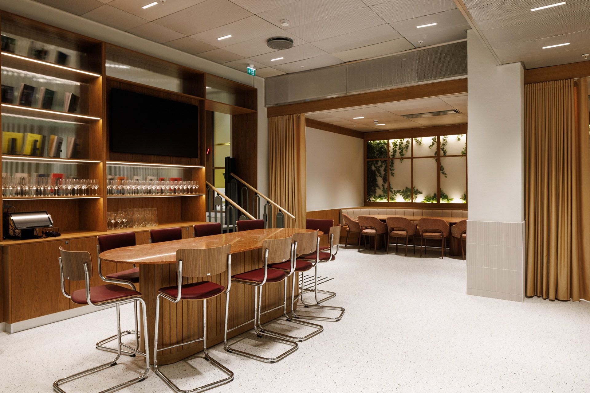 Varm, moderne barlounge med trehyller, høye barkrakker og dempet belysning bak et hvitt gulv.