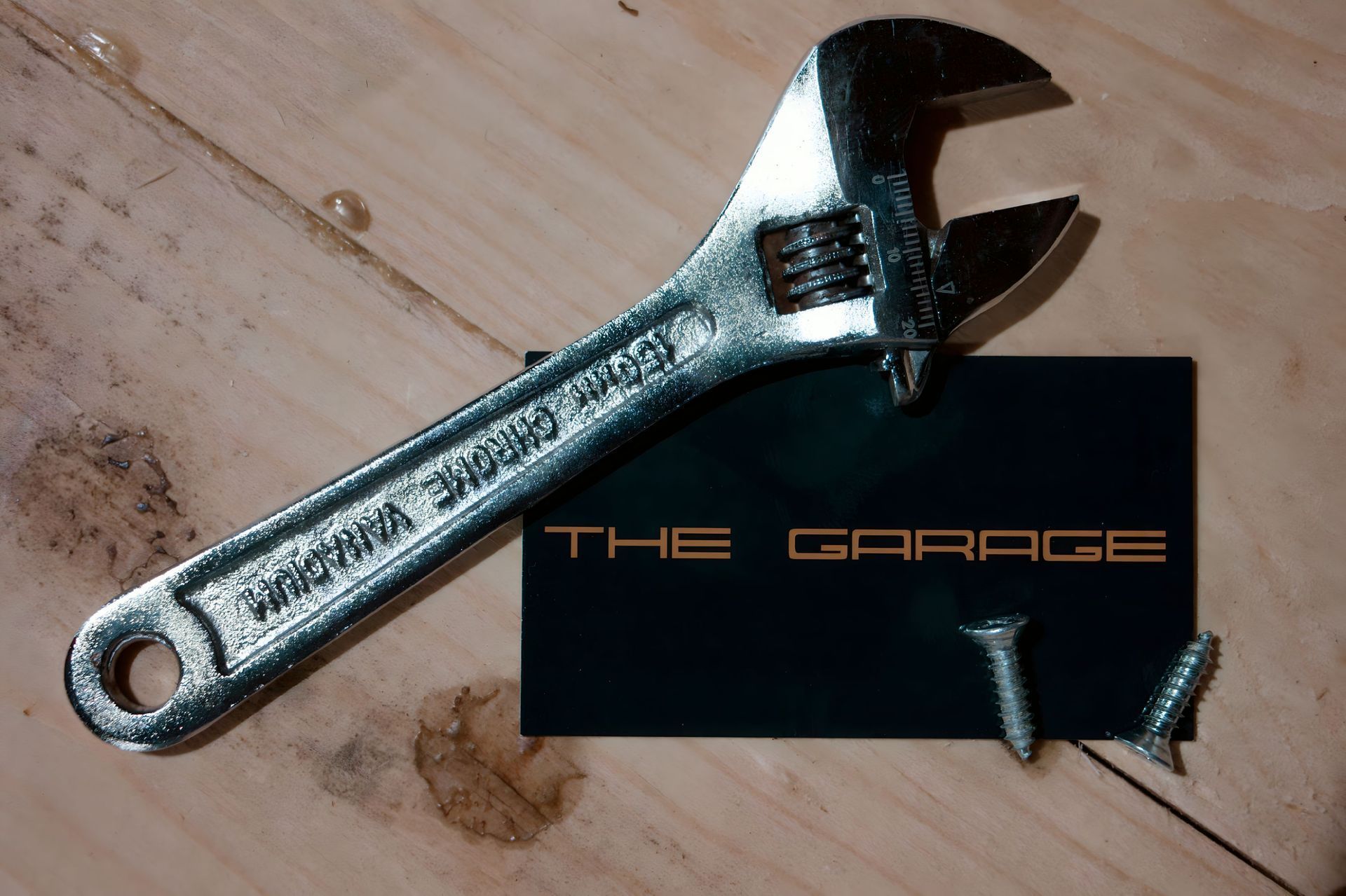 Silver wrench and black card labeled “THE GARAGE” on a wooden surface, with a screw attached | The Garage MKG