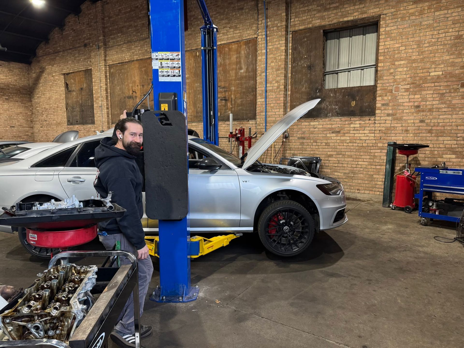 Silver car on a lift in an auto repair shop, with its hood open and a mechanic working nearby. | The Garage MKG