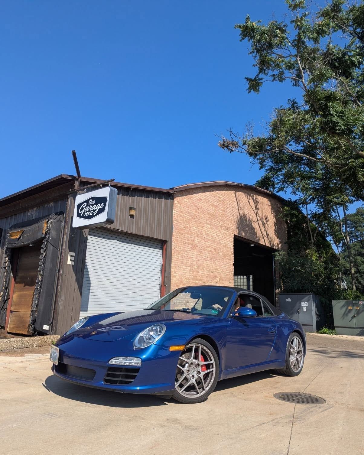 Blue convertible sports car parked outside a small brick auto shop on a sunny day | The Garage MKG