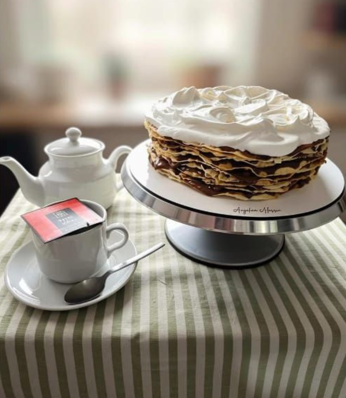 Tarta de crepes apilada con nata montada sobre un soporte, al lado de una tetera y una taza de té, sobre un mantel a rayas.