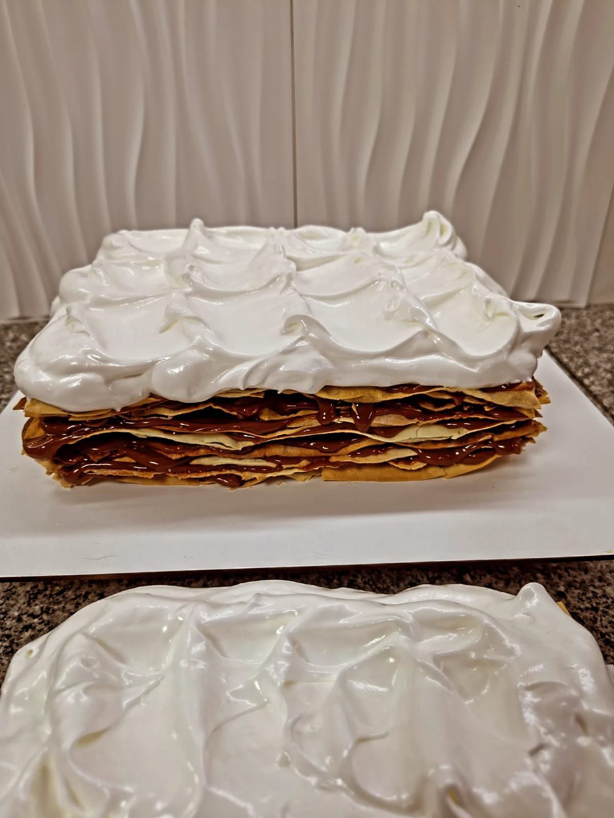 Tarta de crepes apilada con nata montada sobre un soporte, al lado de una tetera y una taza de té, sobre un mantel a rayas.