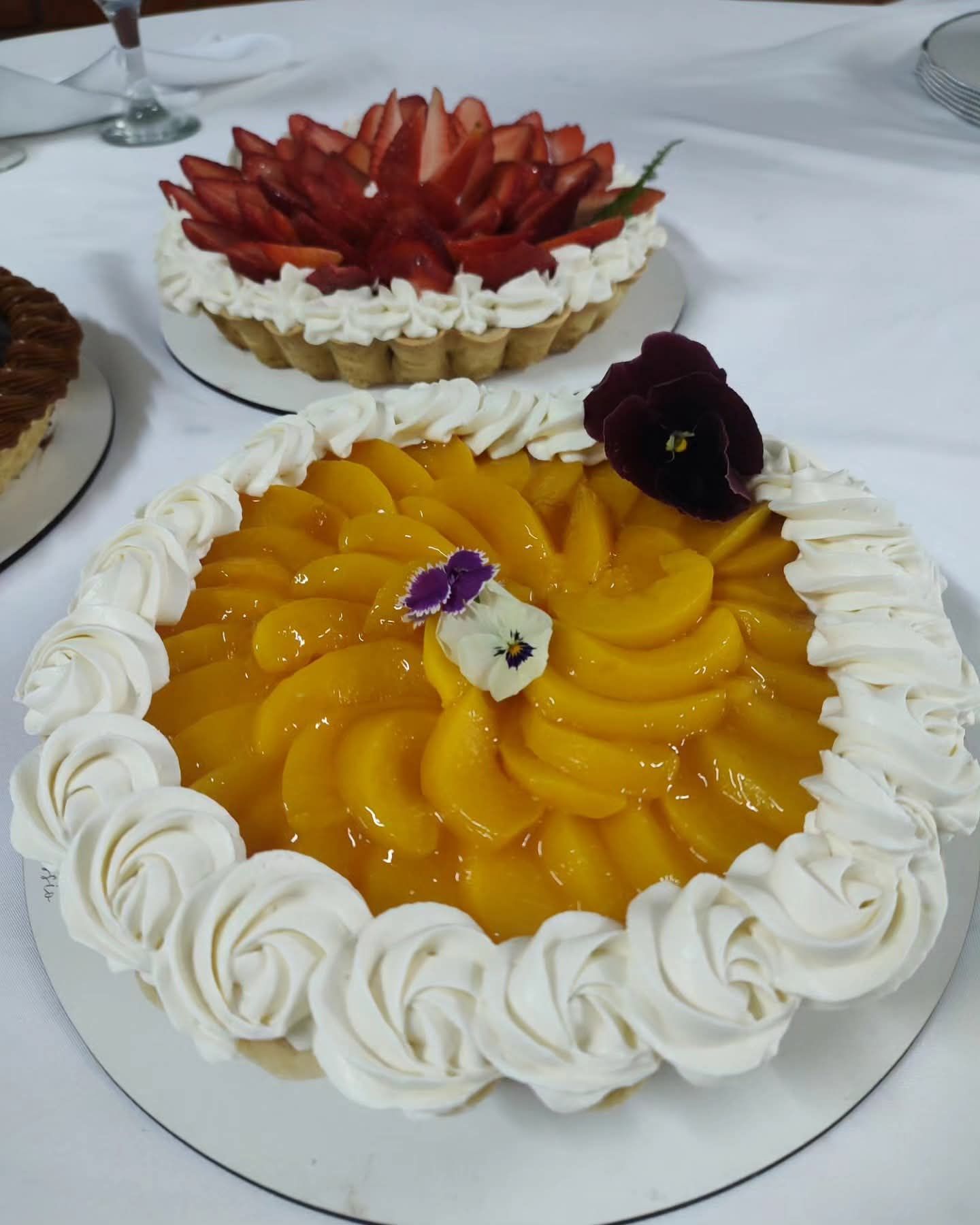 Dos tartas con fruta sobre una mesa: una tarta de melocotón con nata montada y, detrás, una tarta de fresa.