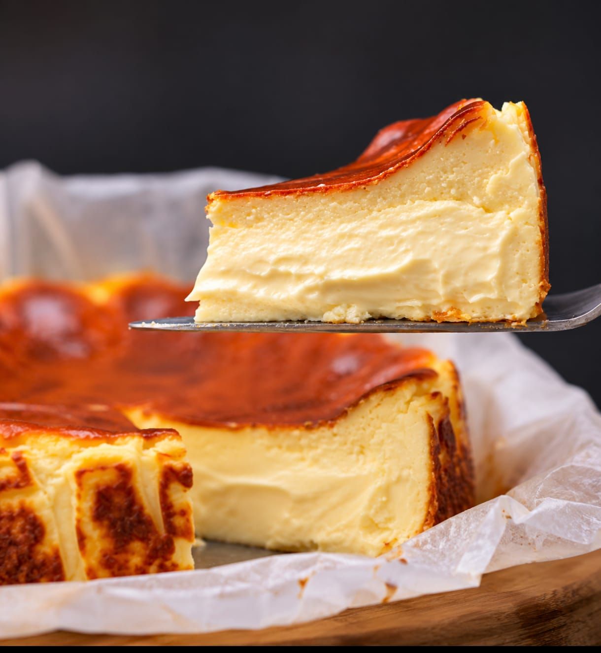 Una porción de tarta de queso vasca quemada sobre una espátula, con más tarta de queso sobre papel de horno al fondo.