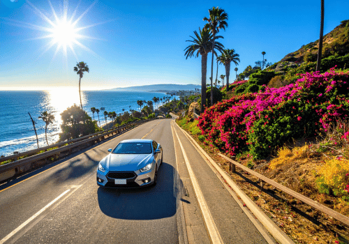 Car driving along a sunny coastal highway lined with palm trees, ocean views, and vibrant hillside.