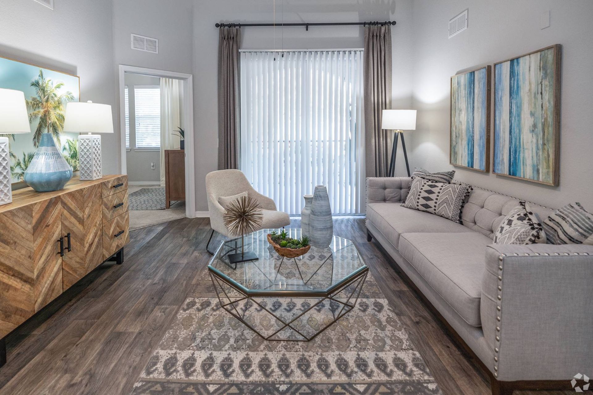 Cozy living room in an apartment with a gray sofa, glass coffee table, and vertical blinds.
