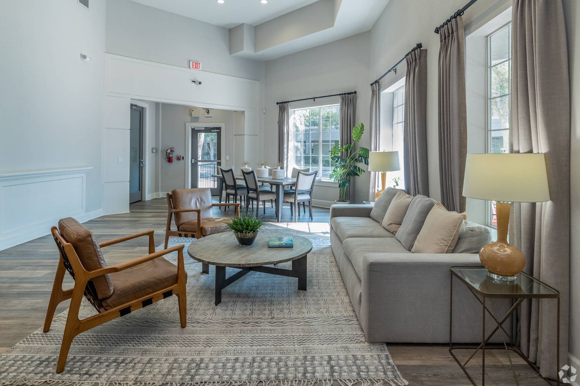 Bright communal living room with sofa, two armchairs, coffee table, dining area, and large windows with curtains.