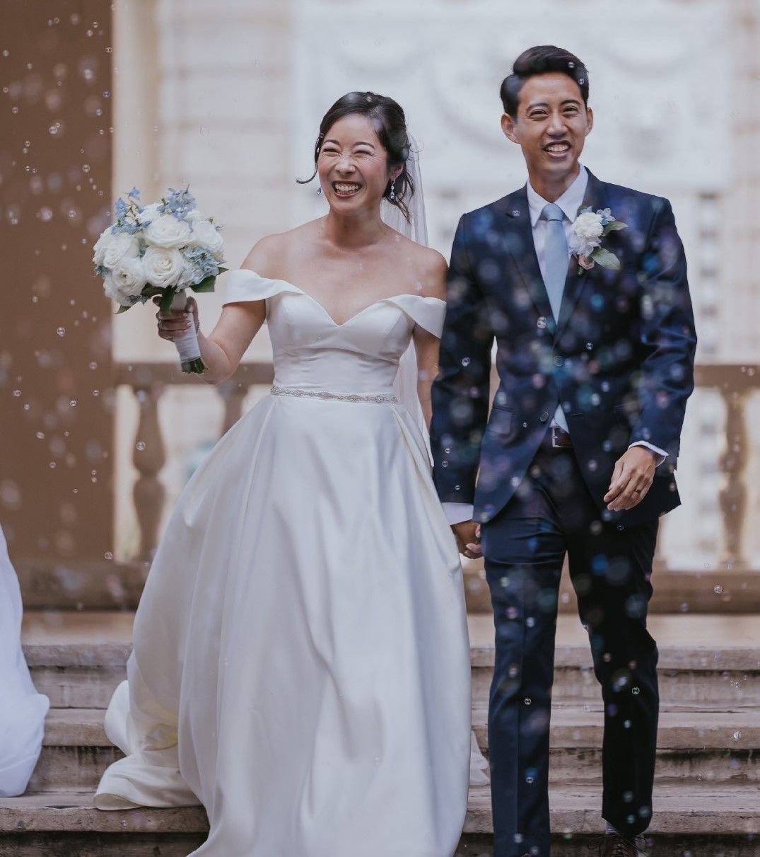 Newlyweds walking together in confetti, bride in white gown holding bouquet, groom in dark suit smiling