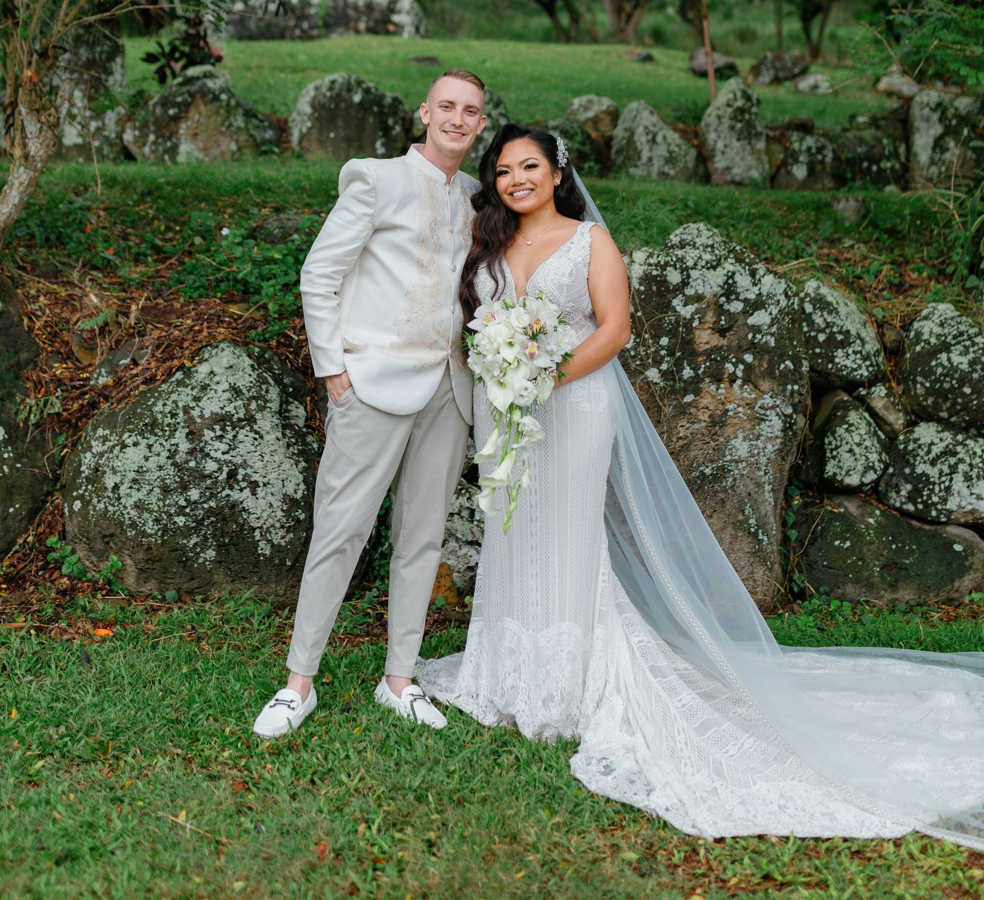 Newlyweds posing outdoors in wedding attire by mossy rocks and greenery, smiling together