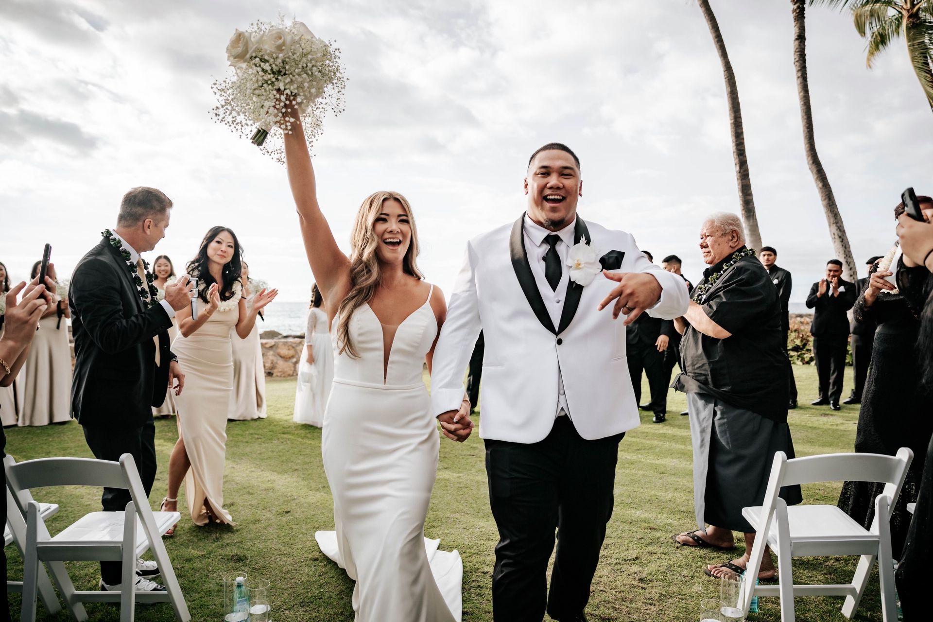 Newlyweds walking down the aisle outdoors, bride holding bouquet aloft, guests cheering on both sides
