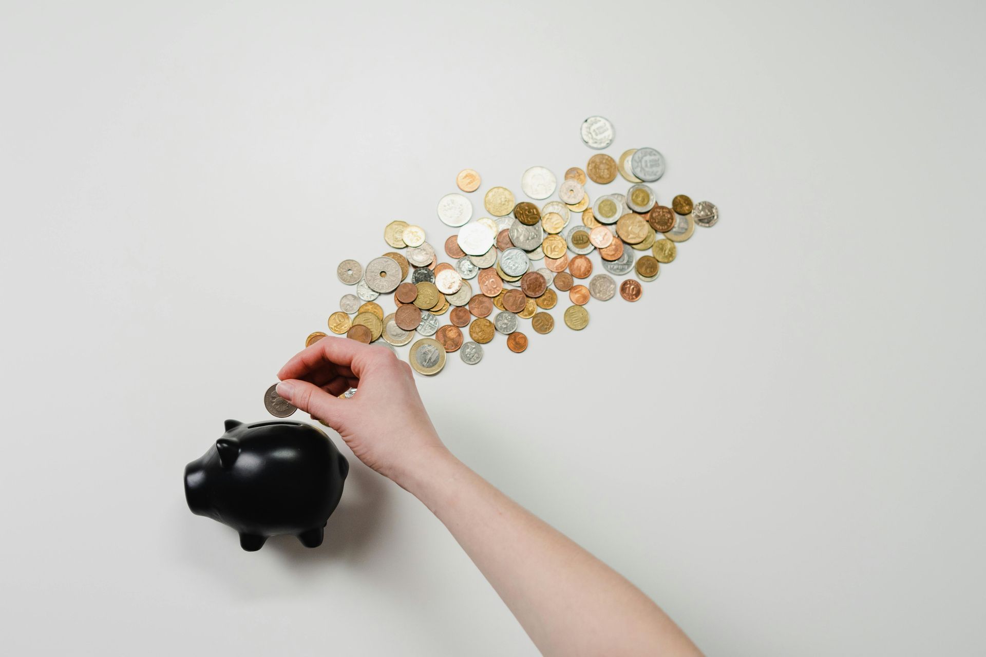 Person putting a coin into a black piggy bank, coins scattered on a white surface.