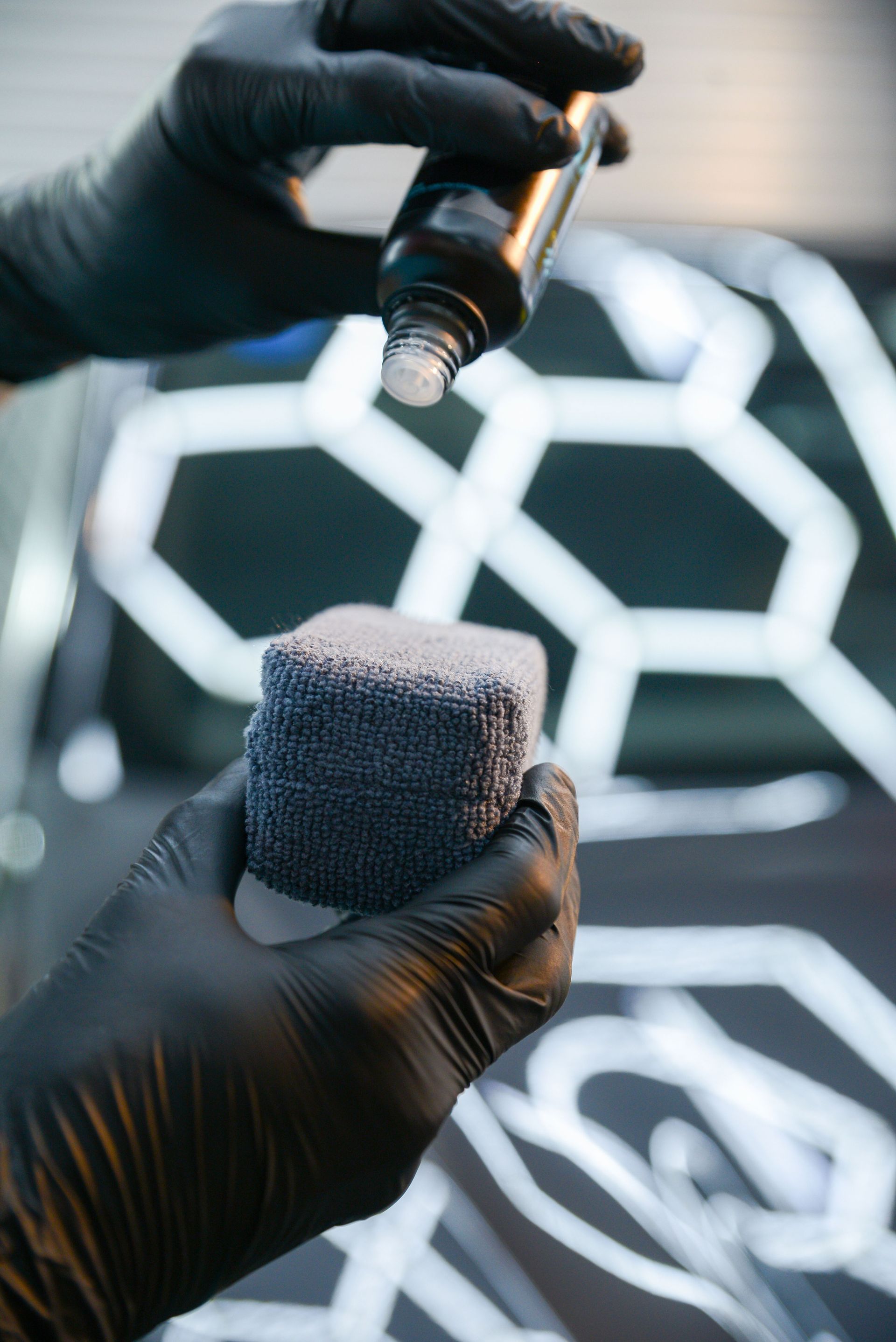 Gloved hands applying liquid from a bottle onto a blue sponge; auto detailing in progress, with car detail lights.