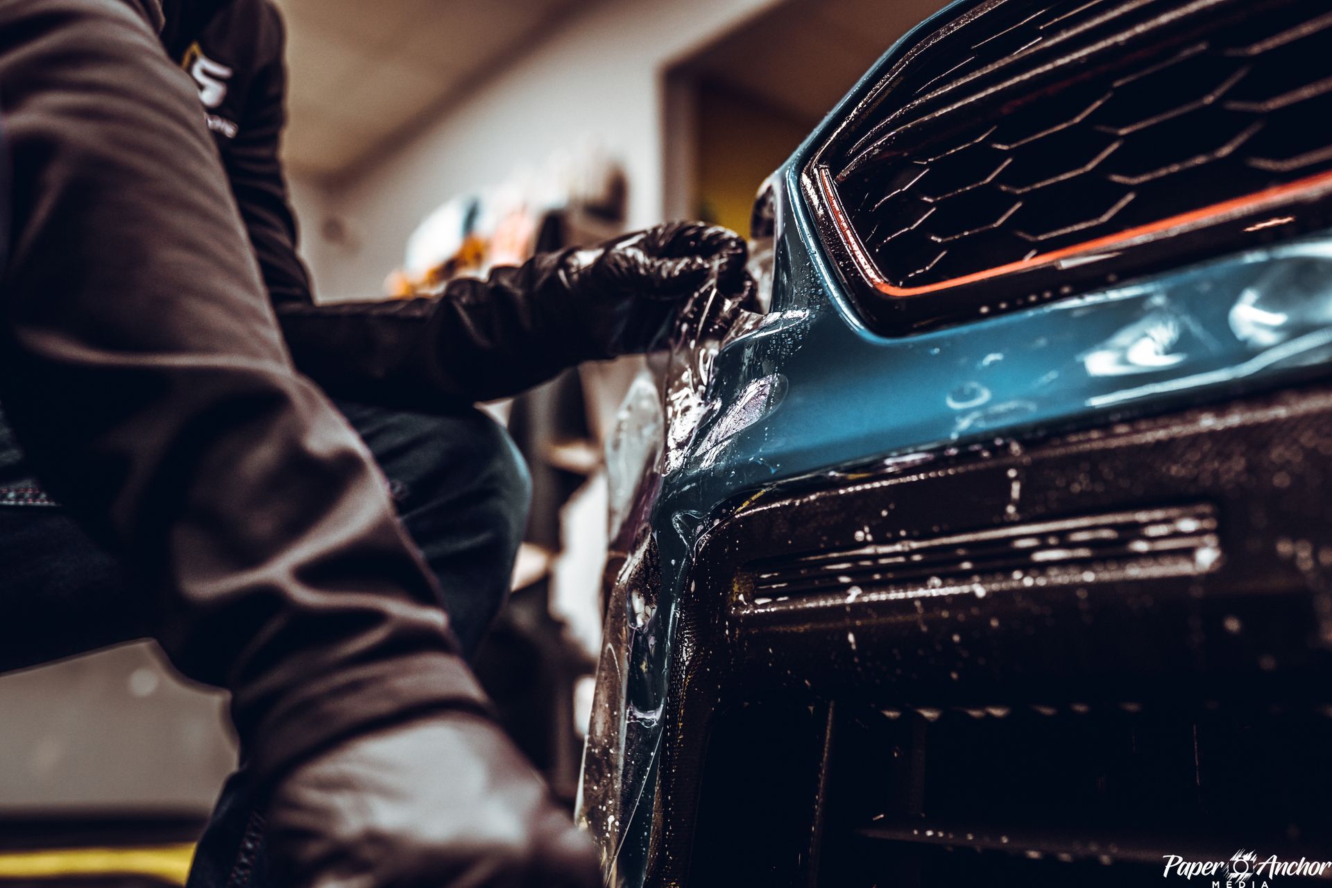 Person washing a blue car, detail shot of the front, suds and a dark-gloved hand.