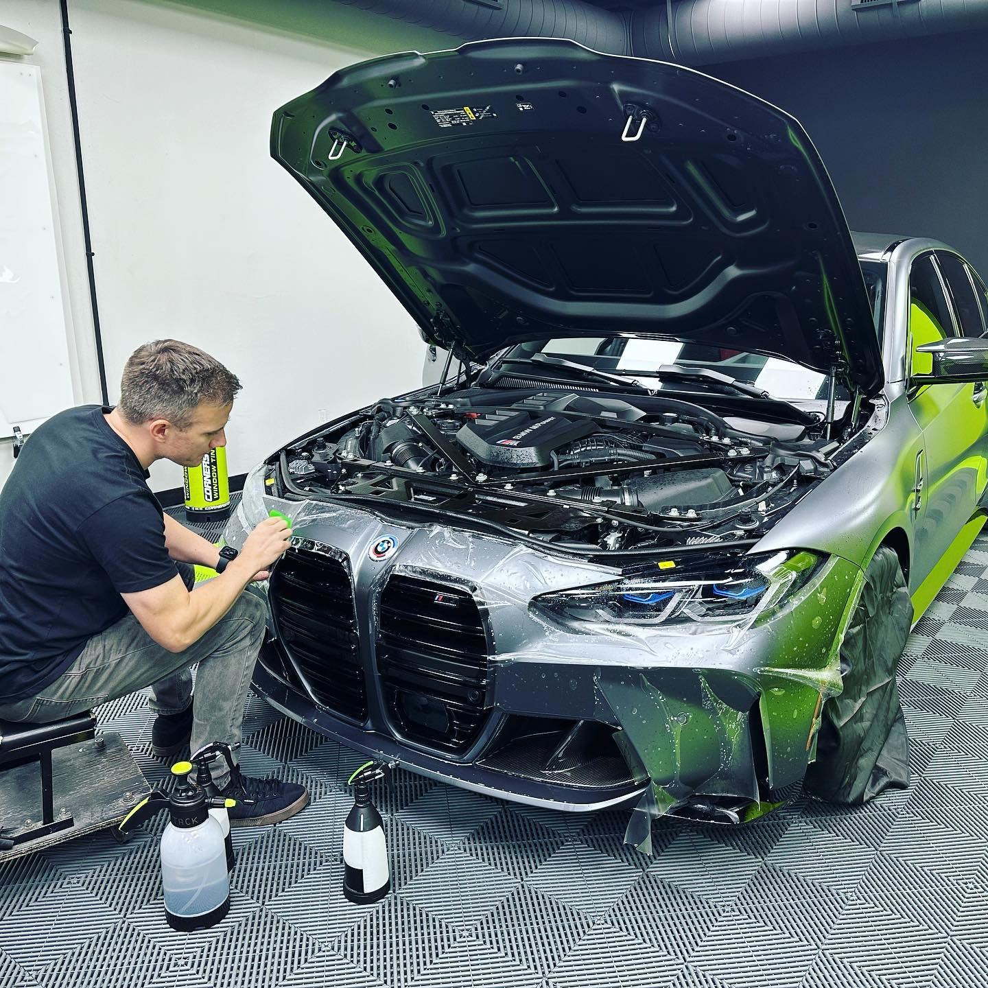 Man working on a gray BMW with the hood open, likely applying a film. Shop setting.