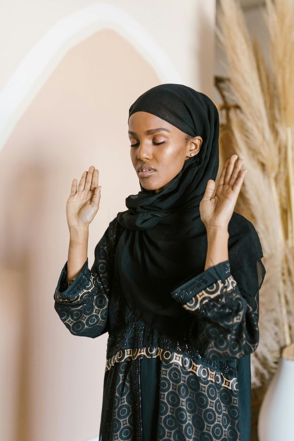 Personne portant un hijab noir et une robe à motifs, les mains levées en prière à l’intérieur