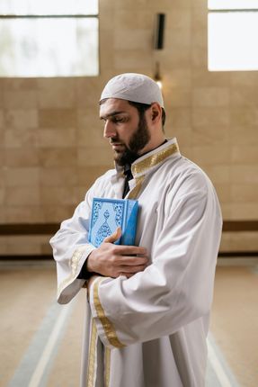 Un homme en robe blanche et portant une calotte tient un livre bleu à l’intérieur d’une mosquée