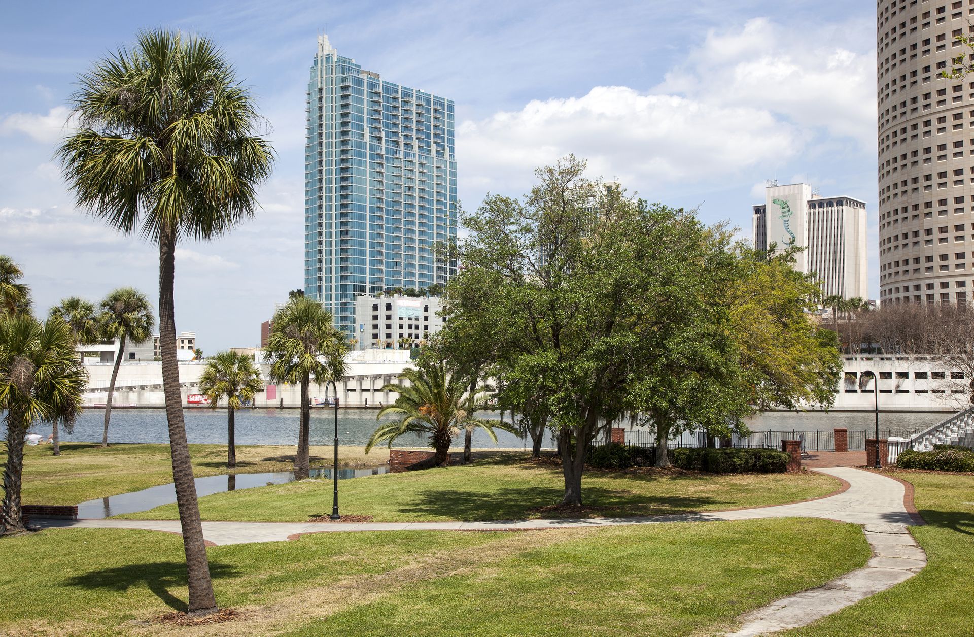 Waterfront park with palm trees, winding path, and tall city buildings under a partly cloudy sky