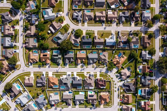 Aerial view of a suburban neighborhood with rows of houses, streets, trees, and parked cars.