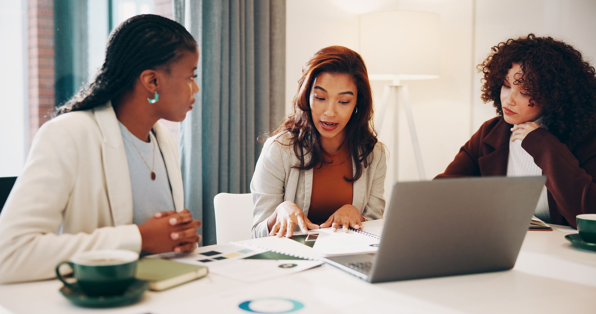Three coworkers discuss documents at a table with a laptop in a bright office