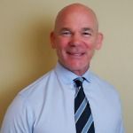 A bald man wearing a blue shirt and tie is smiling for the camera.
