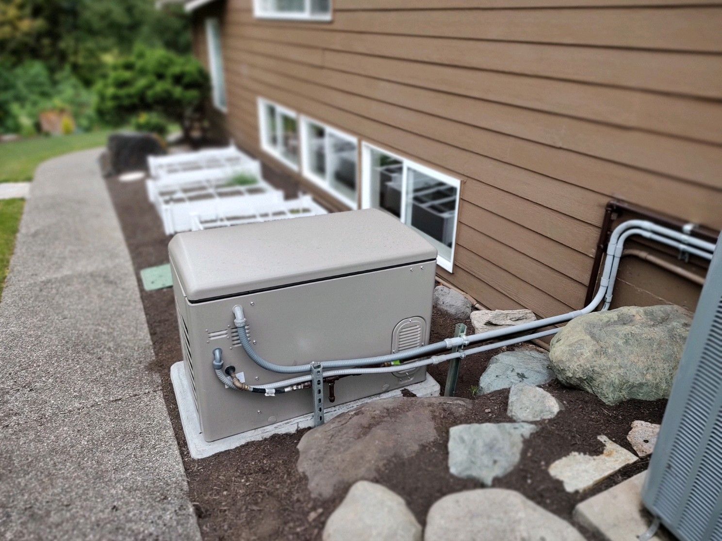 A brown generator next to a house with large rocks and a garden bed.