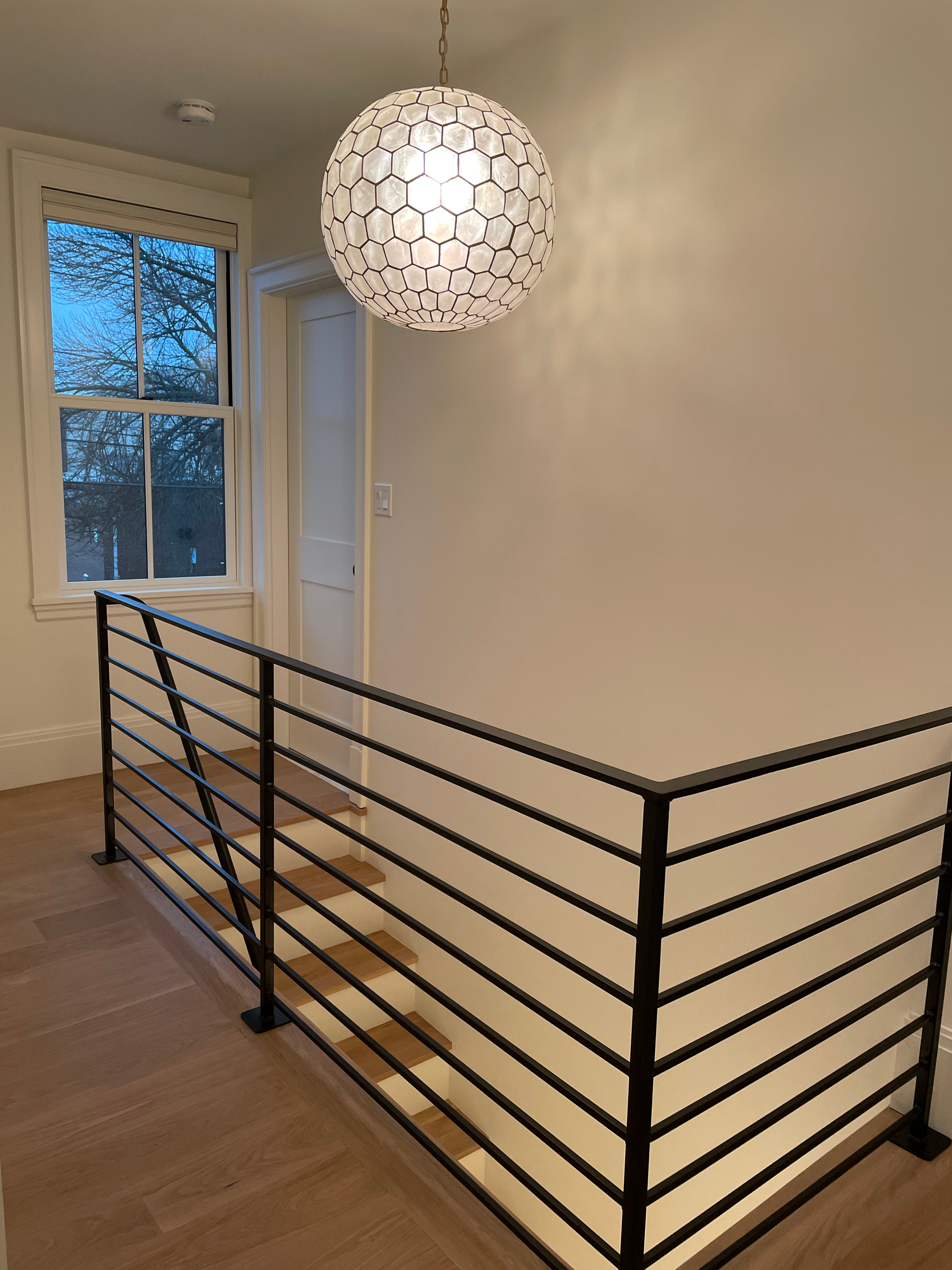 Modern staircase landing with black railing, wooden steps, and a hanging white pendant light near a window.