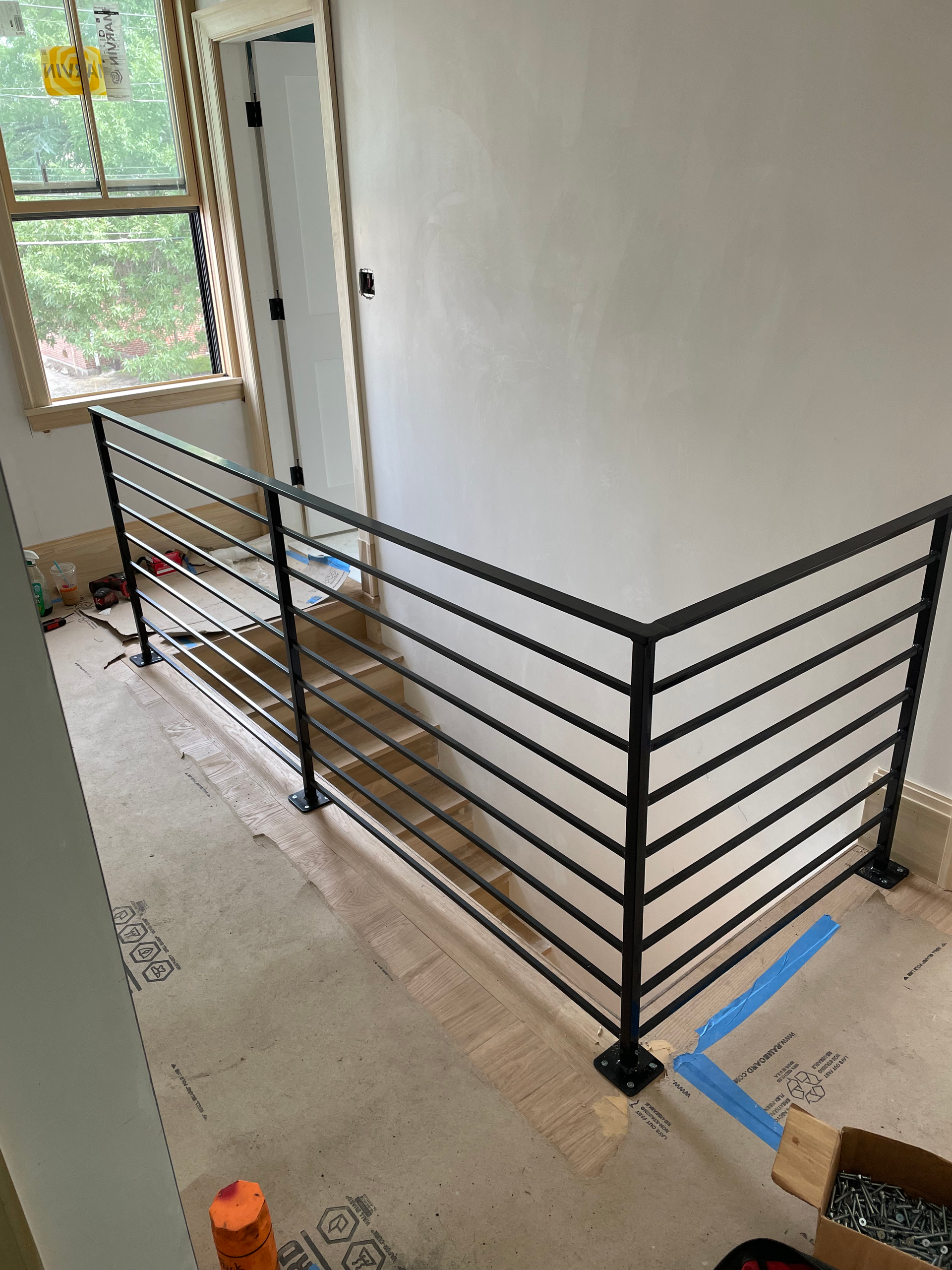 Stair landing with black metal railing beside a white wall and window; unfinished floor and taped-off area.
