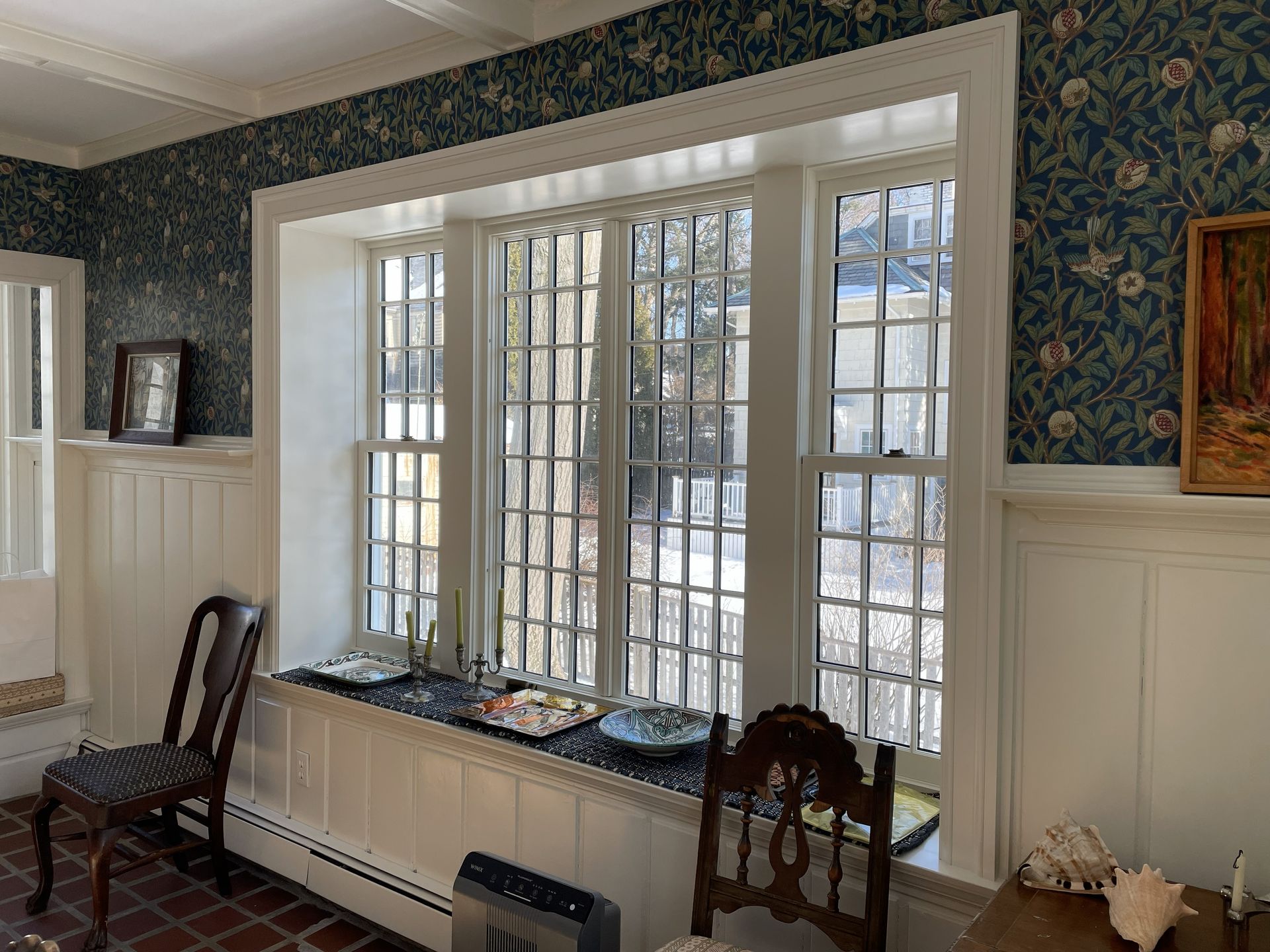 Bright sunroom with large white-paned windows, patterned wallpaper, wainscoting, and wooden chairs.