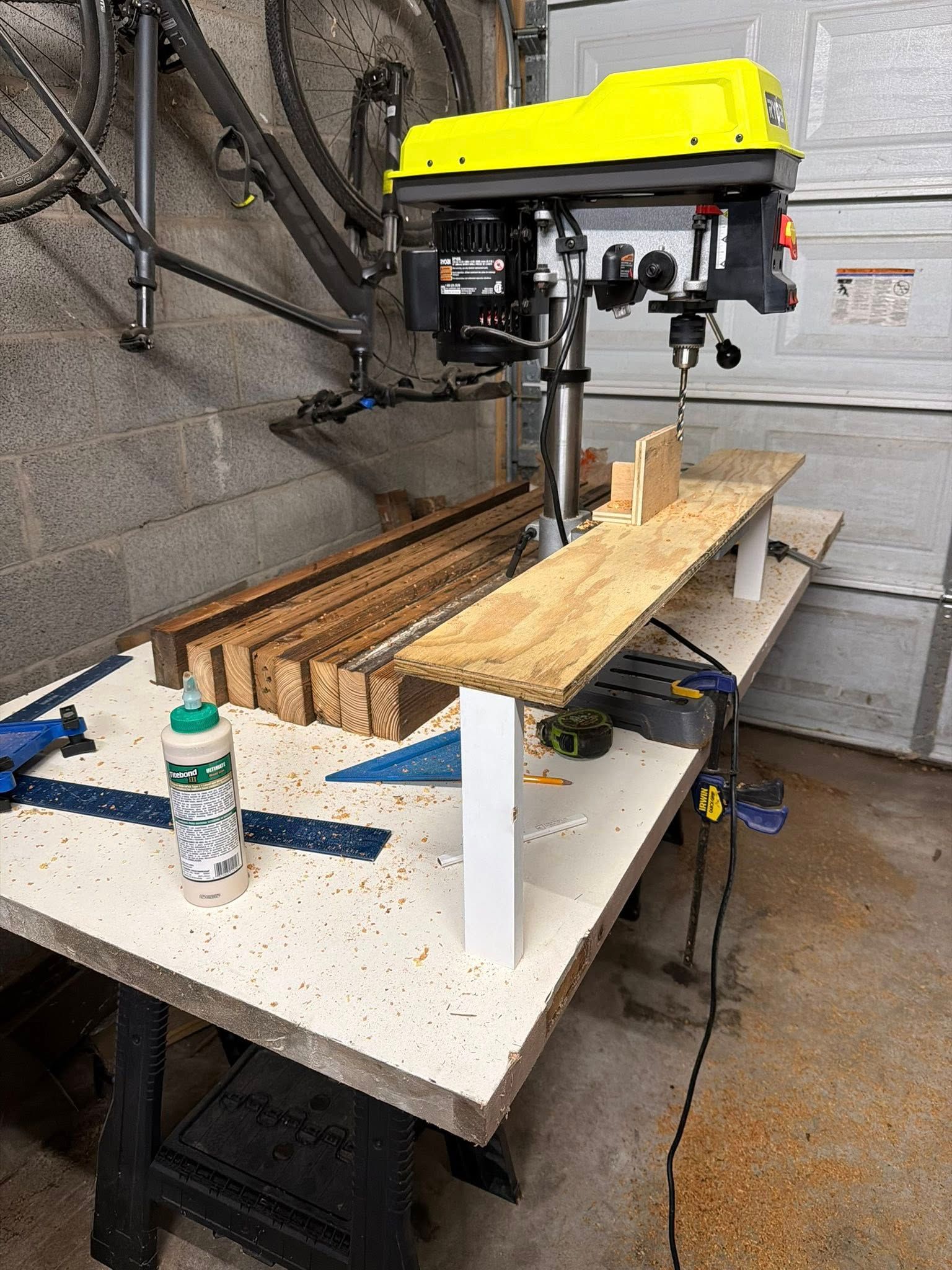 Yellow drill press over a workbench with wood pieces and tools in a workshop