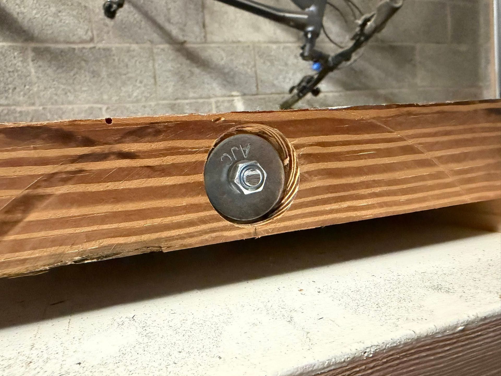 Close-up of a metal bolt securing a chain to a wood board against a concrete wall.