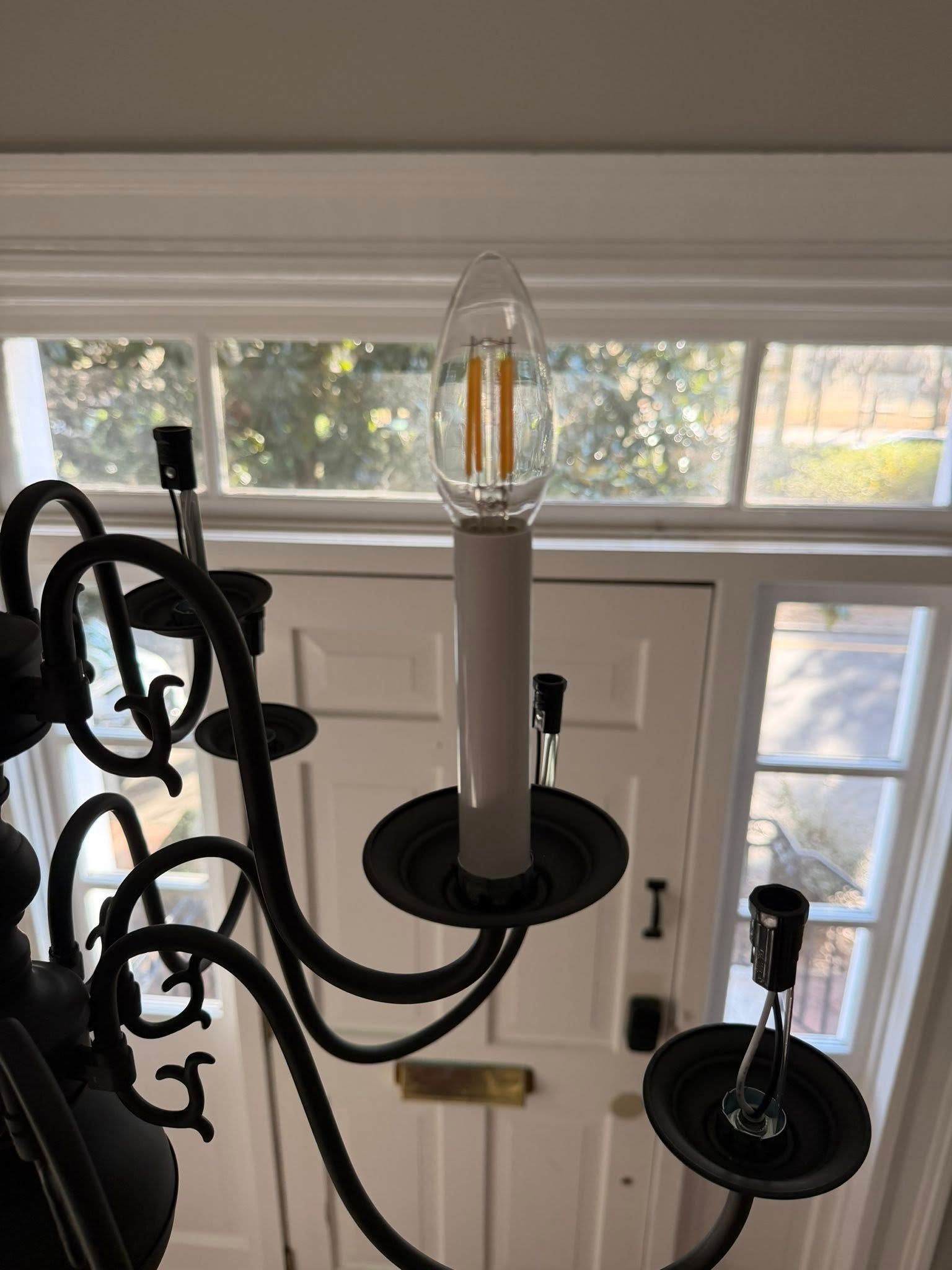 Black chandelier with candle-style bulbs hanging by a bright window and front door.