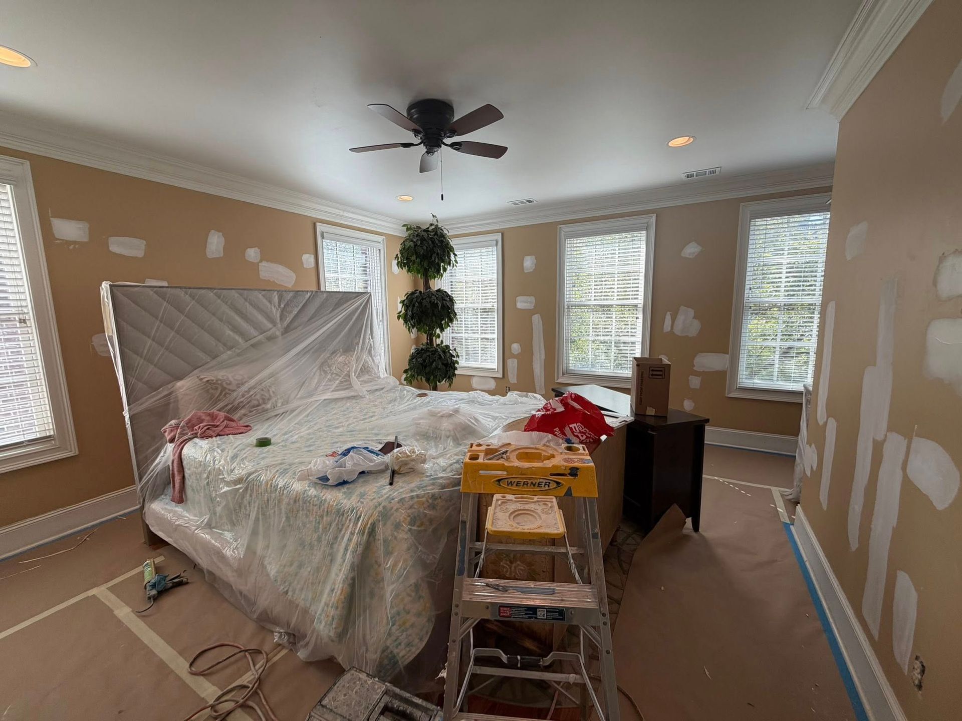 Bedroom under renovation with taped walls, covered bed, ladder, and paint supplies