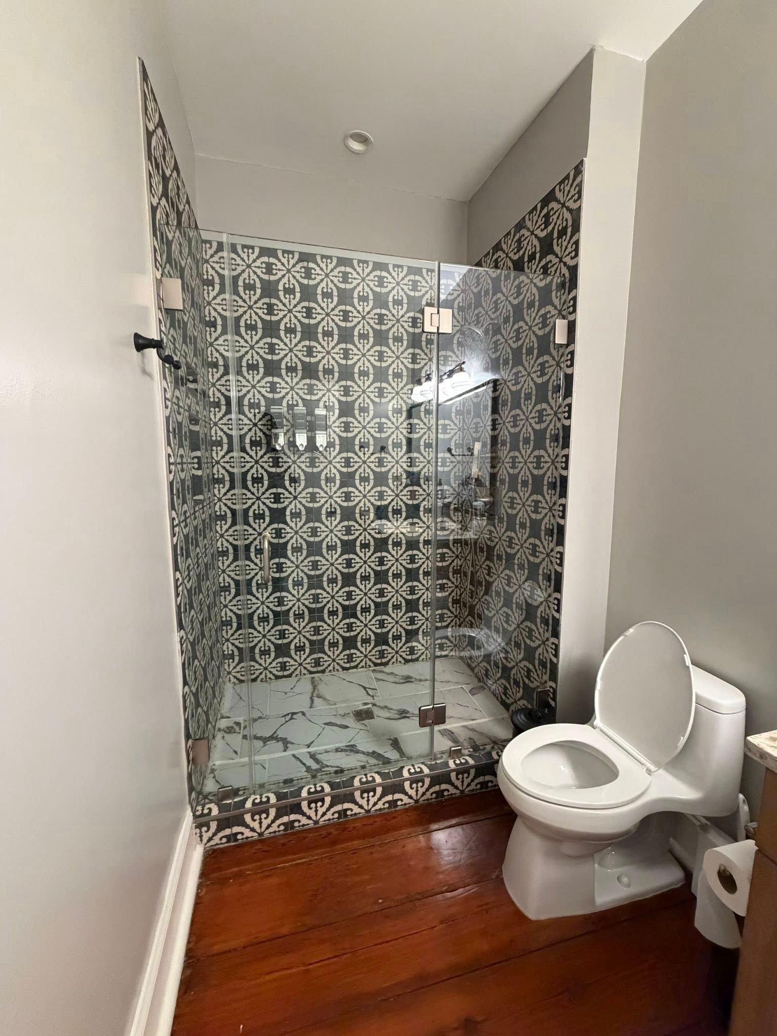 Narrow bathroom with tiled walk-in shower and toilet beside a wood floor.