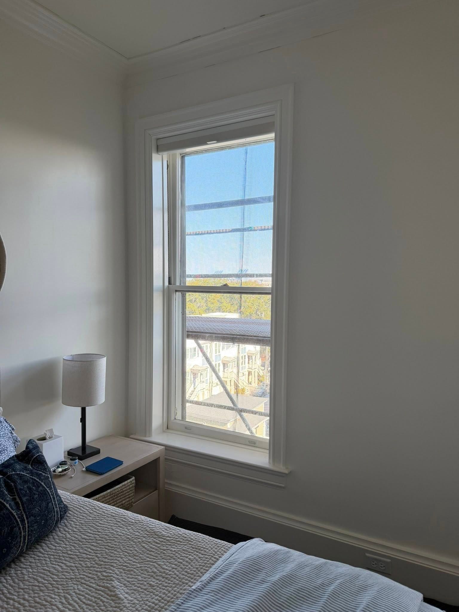 Bedroom corner with bed, nightstand lamp, and a tall window with sunlight and a view outside