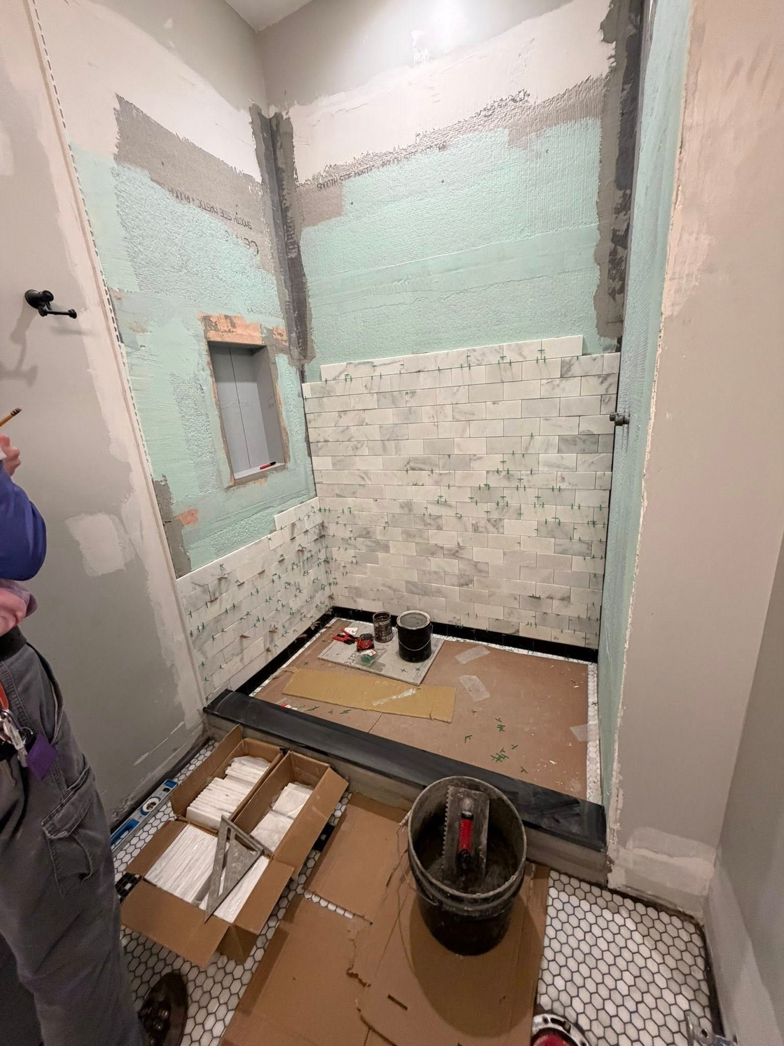 Small bathroom under renovation with tiled shower wall, exposed floor, tools, and construction materials.