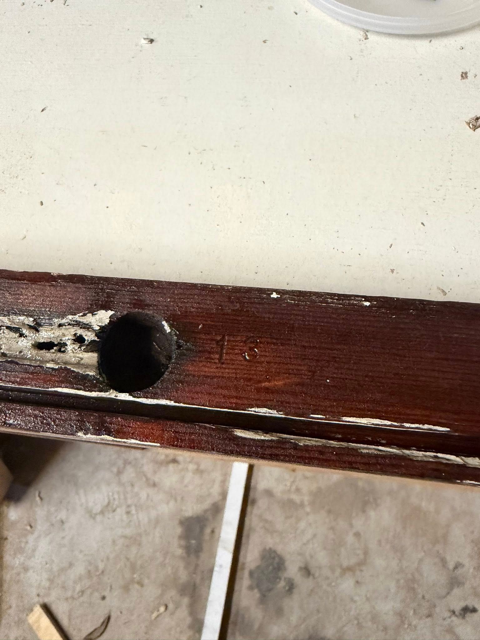 Close-up of a weathered dark wooden board with a drilled hole beside a white wall and concrete floor.