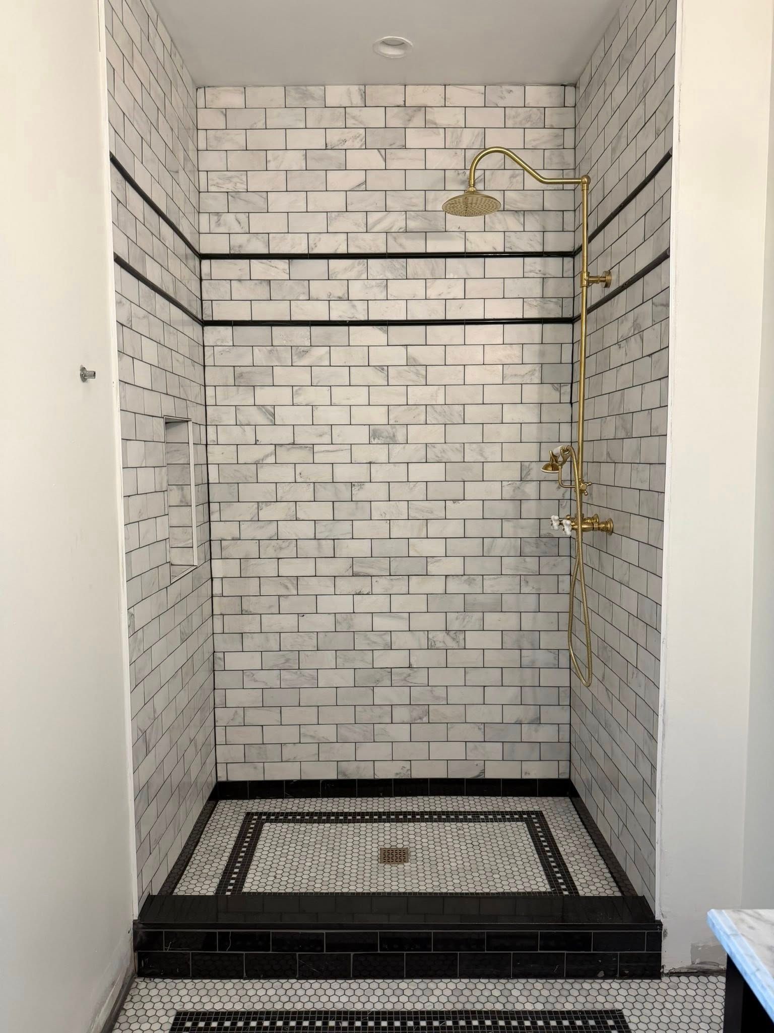 White tiled shower with brass fixtures, black accent border, and mosaic floor in a narrow bathroom alcove