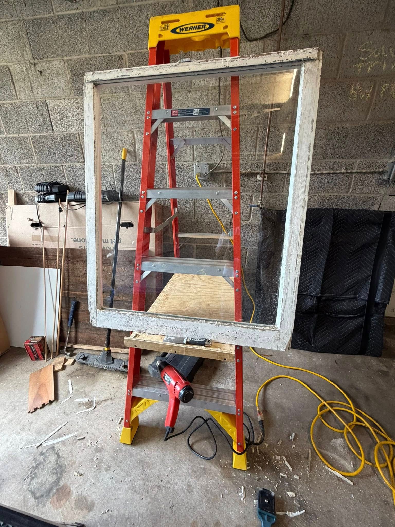 A red ladder behind a white foam-framed window mockup in an unfinished workshop with cables on the floor