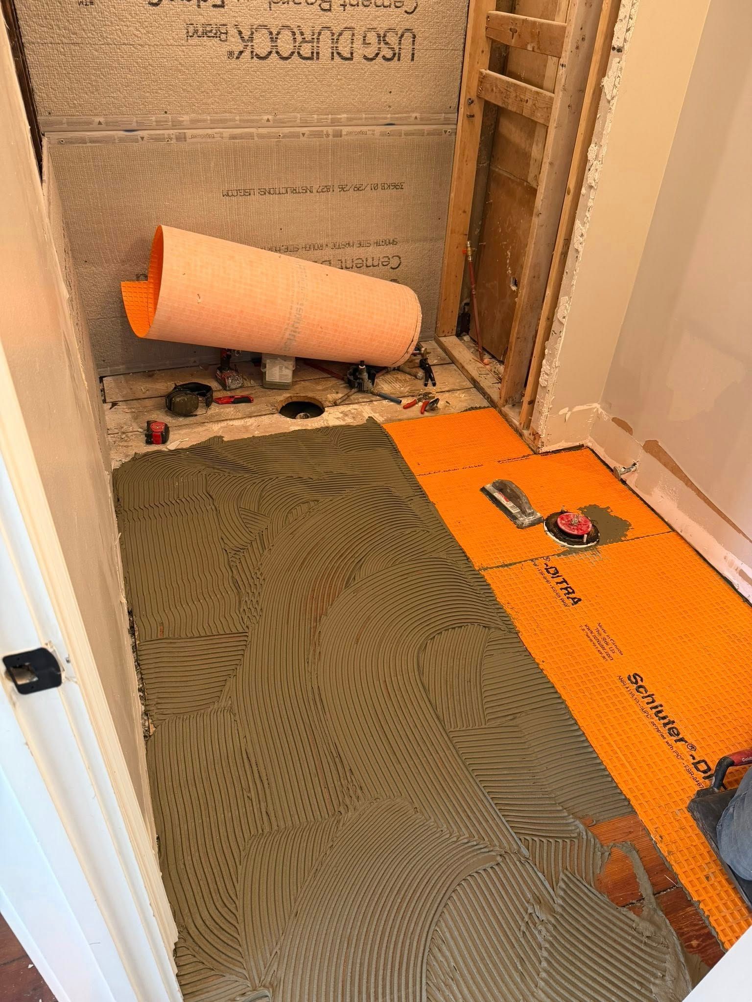 Partially tiled shower floor with orange underlayment, rolled membrane, and tools during renovation